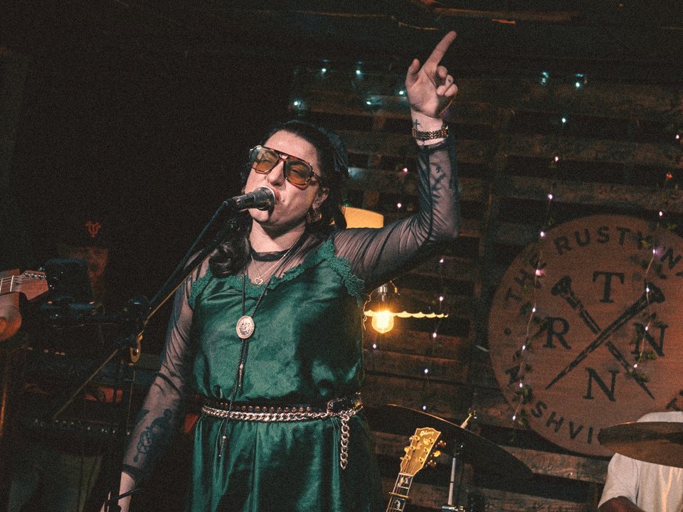 Tori singing with her hand raised in front of The Rusty Nail sign